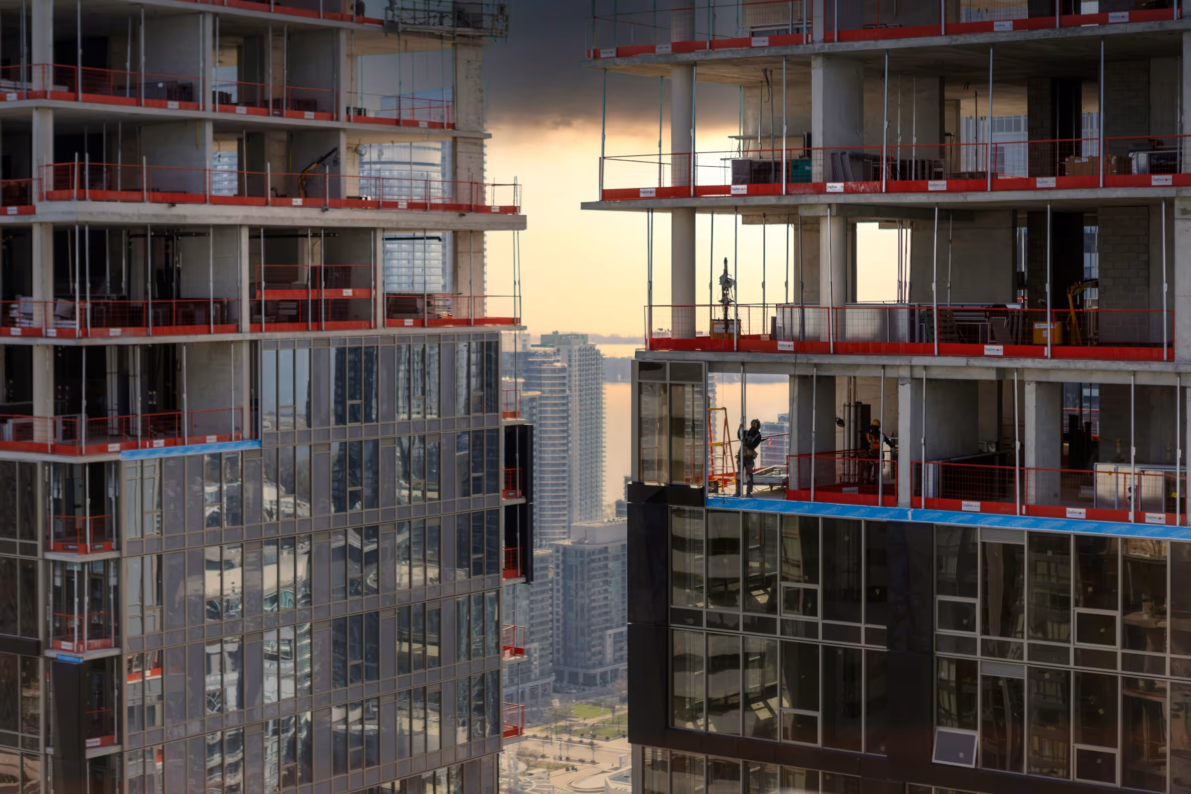 Two high-rise buildings under construction with workers on the top floors and a cityscape under a cloudy sky in the background.