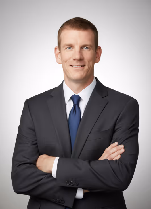 Pascal Monat in a dark suit, light blue shirt, and maroon tie standing with arms crossed, smiling against a gray background.