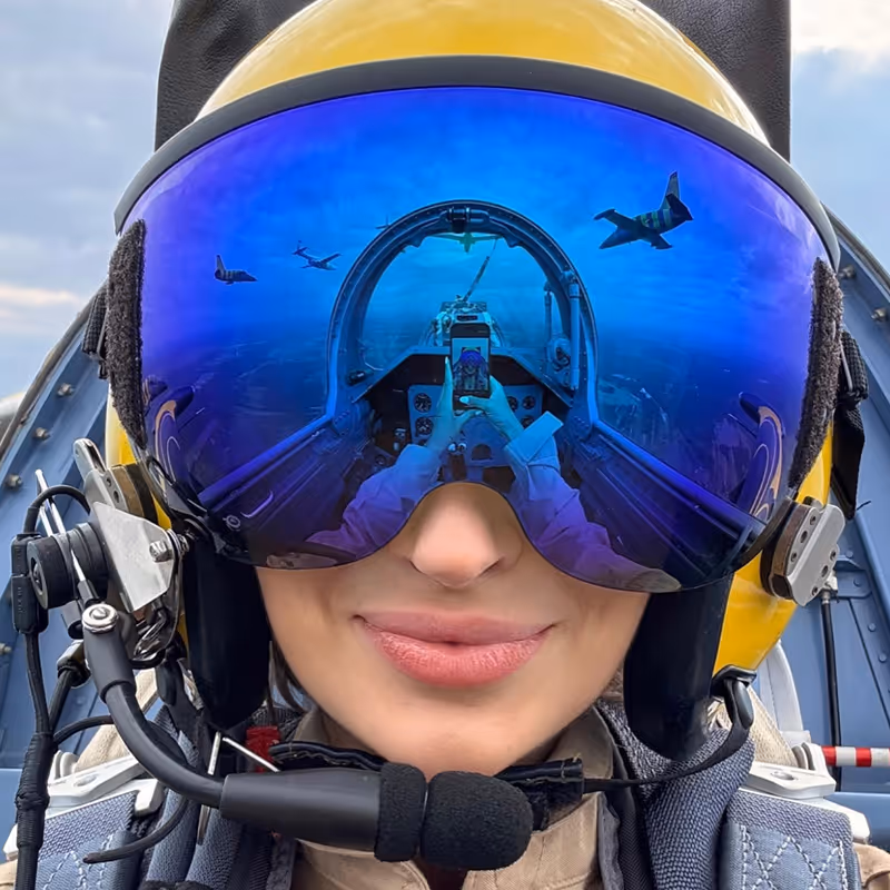 Close-up of a female pilot wearing a yellow helmet and reflective blue visor showing aircraft cockpit and fighter jets in the reflection.