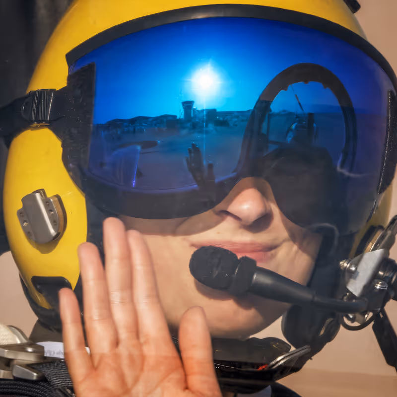 Close-up of a person wearing a yellow helmet with blue reflective visor and a microphone, raising their hand in greeting.