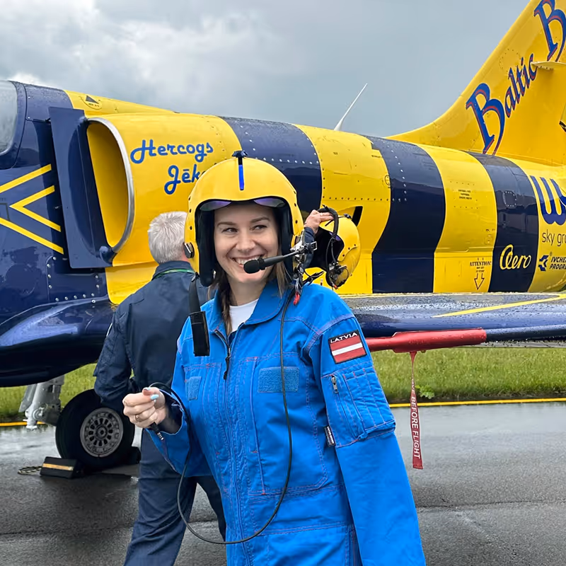Smiling female pilot wearing a blue flight suit with a Latvia patch and a yellow helmet standing in front of a yellow and blue aircraft.