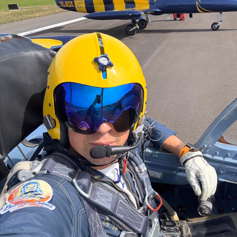 Pilot wearing a yellow helmet with blue reflective visor and flight suit inside a cockpit, holding controls on an airfield.