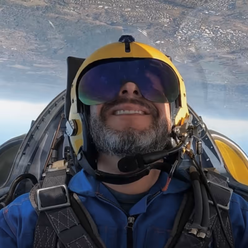 Pilot wearing a yellow helmet and blue flight suit smiling inside a jet cockpit with inverted aerial view outside.