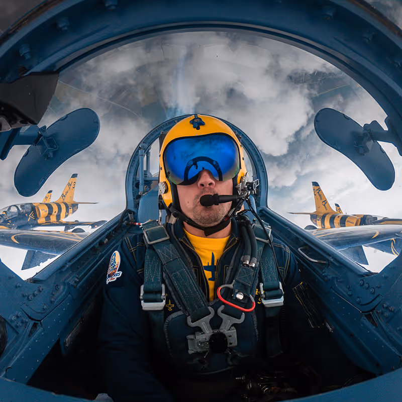 Pilot wearing a yellow helmet and blue reflective visor inside a jet cockpit with two yellow and black striped fighter jets flying in formation against a cloudy sky.