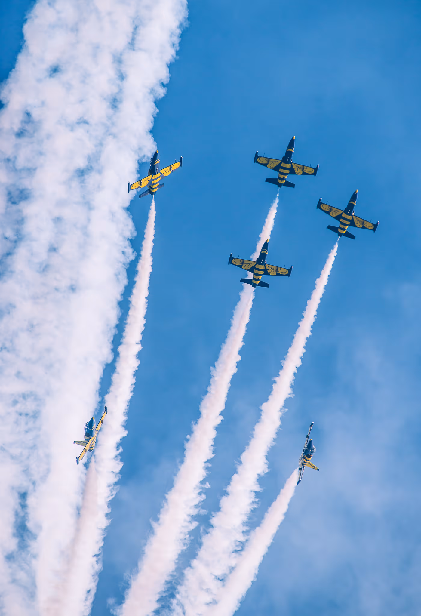 Four airplanes performing a synchronized air show with smoke trails above three sports cars on a runway.