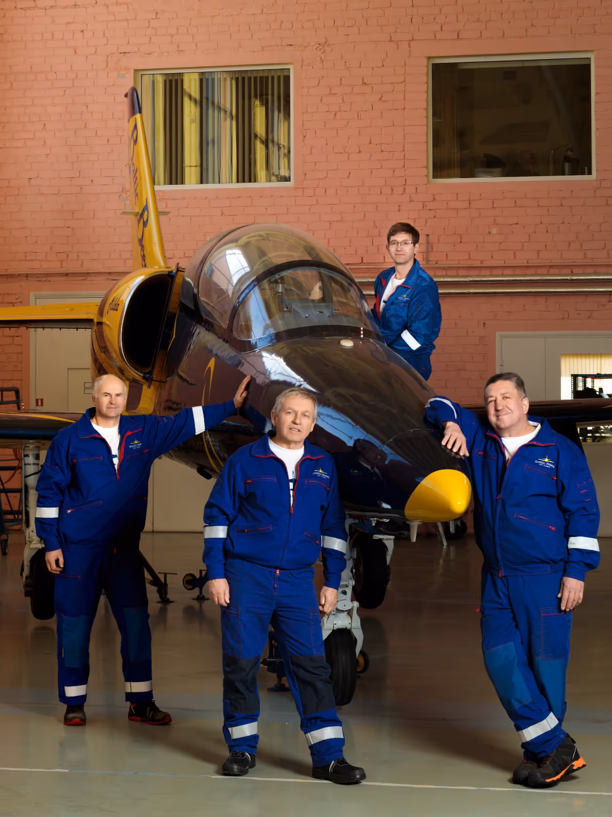 Four men in blue mechanic jumpsuits posing around and on a small jet aircraft inside a hangar.