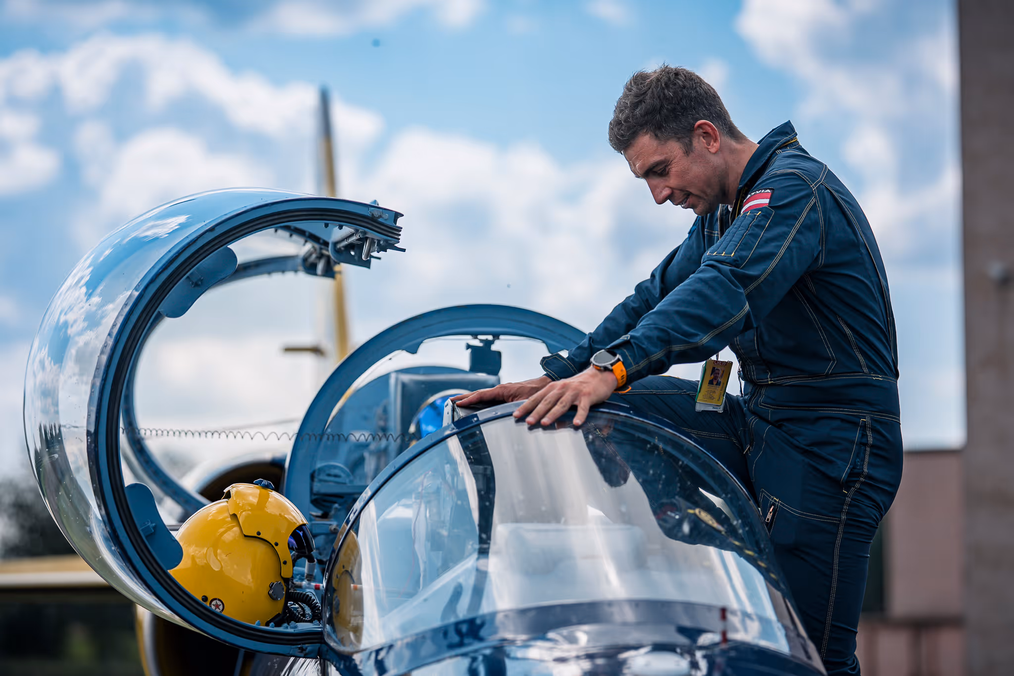 Pilot in a dark blue flight suit preparing a jet cockpit with a yellow helmet inside under a partly cloudy sky.