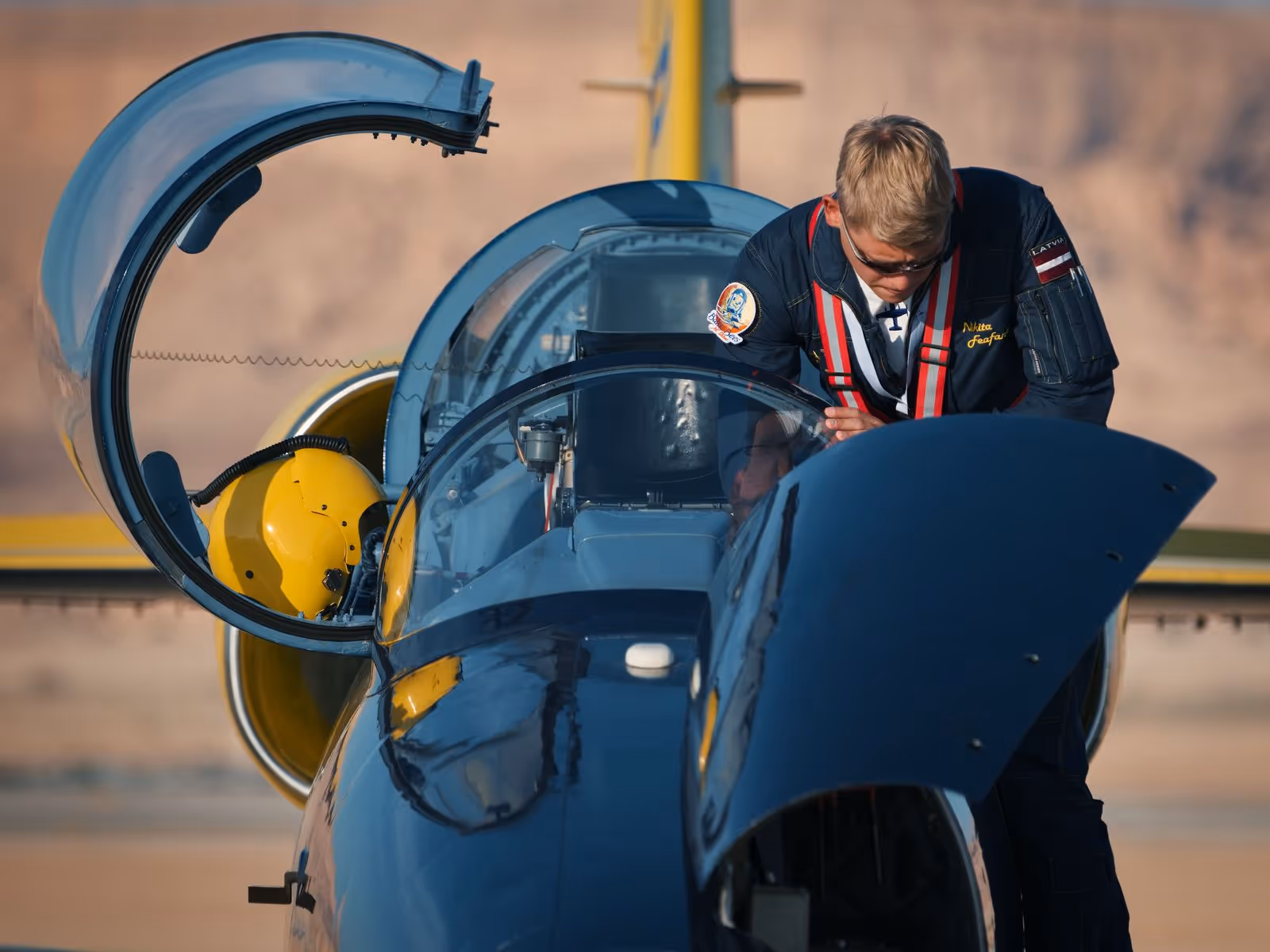 Pilot in flight suit inspecting the cockpit of a blue and yellow fighter jet with the canopy open.