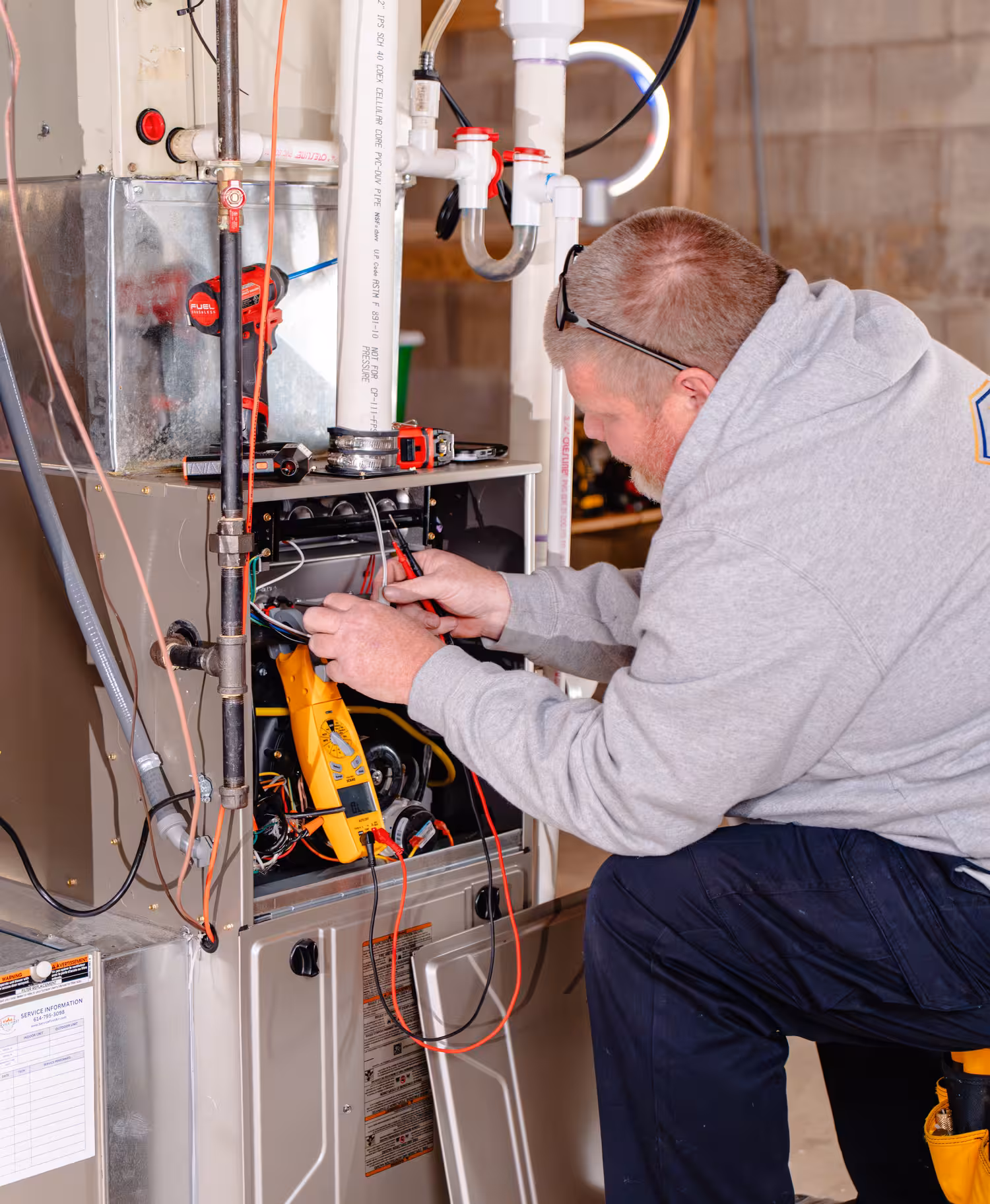 Technician in a gray hoodie using a multimeter to test wiring inside a furnace unit in a basement.