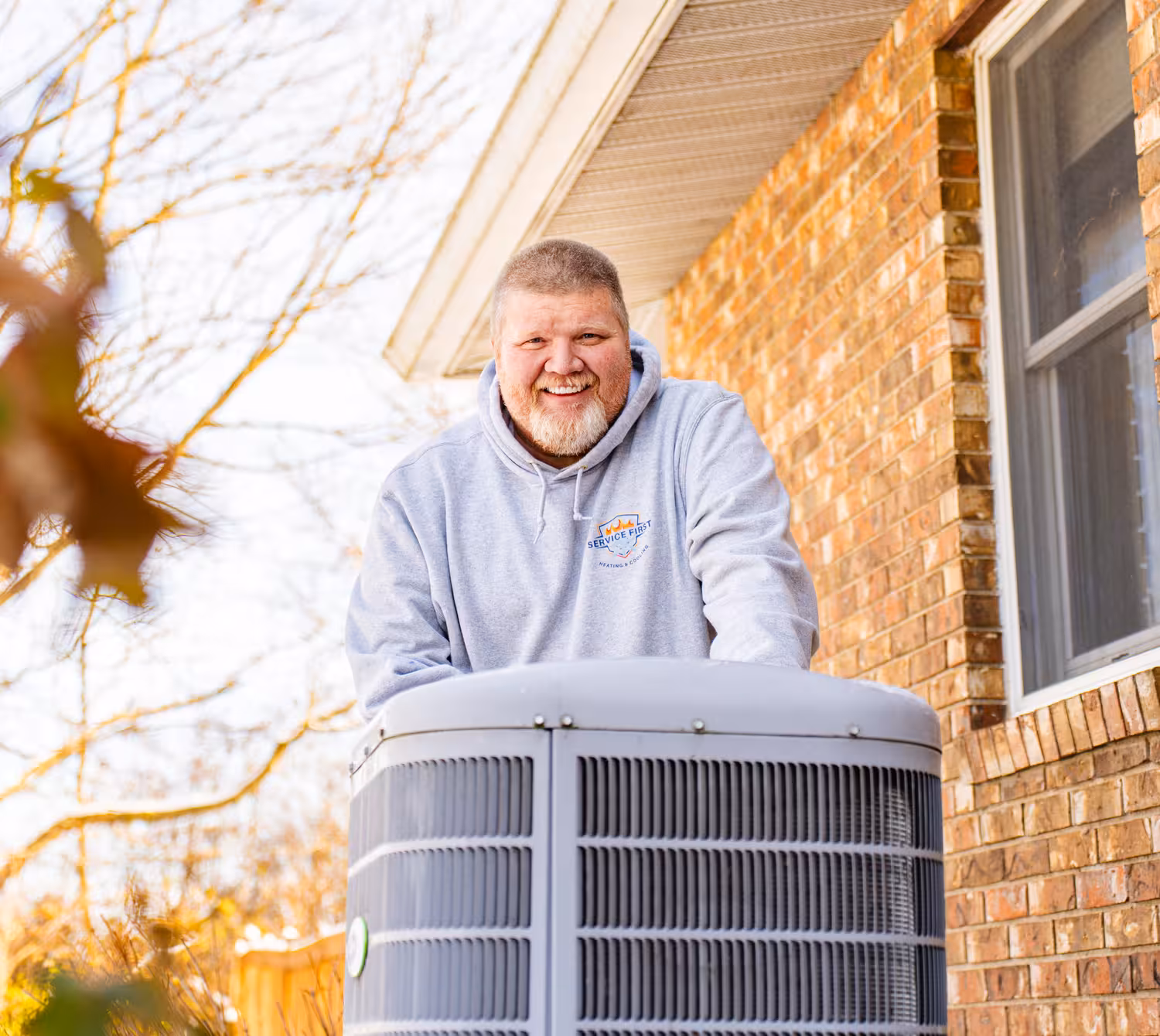 Smiling technician in a gray hoodie inspecting an outdoor air conditioning unit next to a brick house.