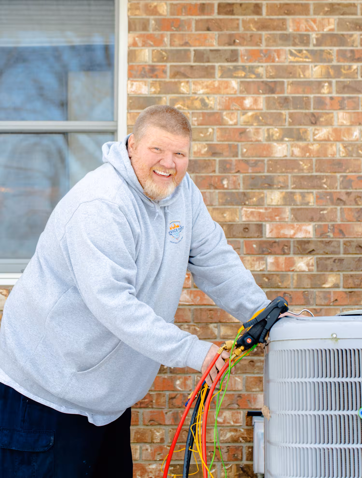Technician in gray hoodie smiling while servicing outdoor air conditioning unit with colorful diagnostic hoses.