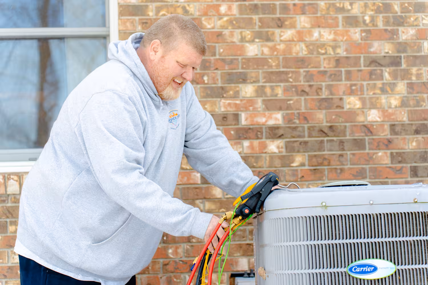 Technician in a gray hoodie inspecting a Carrier heat pump unit outside a brick building.