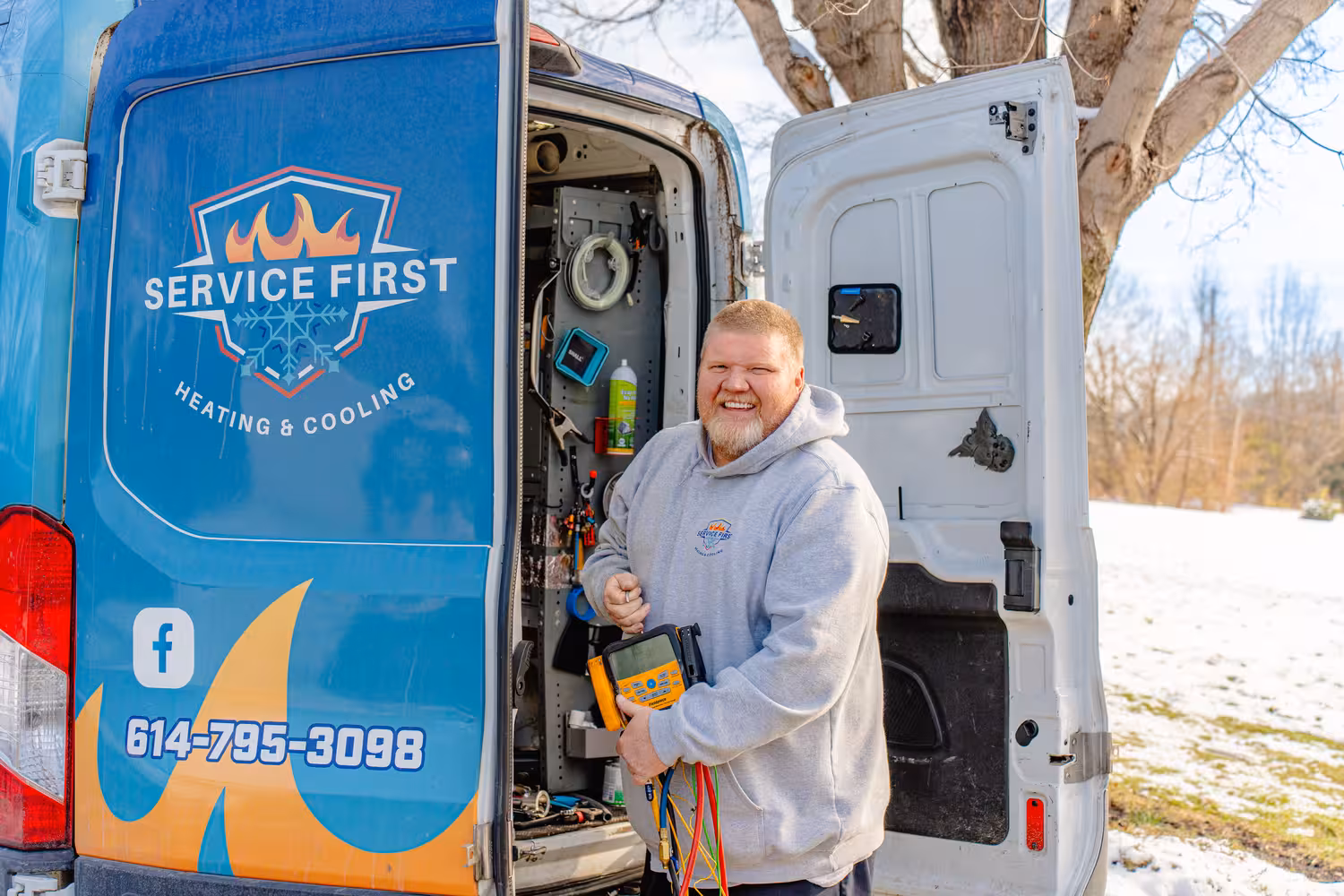 Smiling technician in gray hoodie holding diagnostic equipment for a heat pump standing beside an open service van with Service First Heating & Cooling branding.