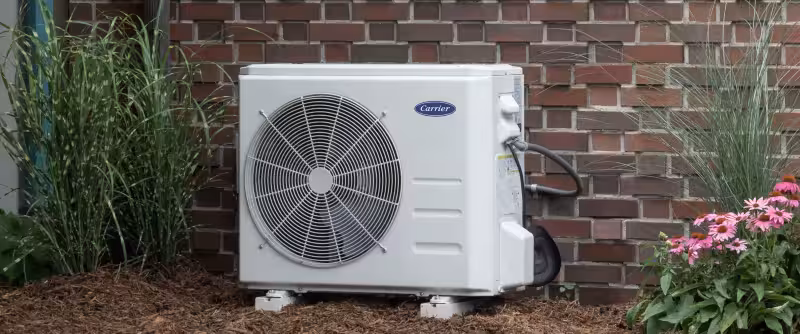 Carrier brand outdoor mini-split air conditioner unit installed on mulch against a brick wall with green plants on the sides.