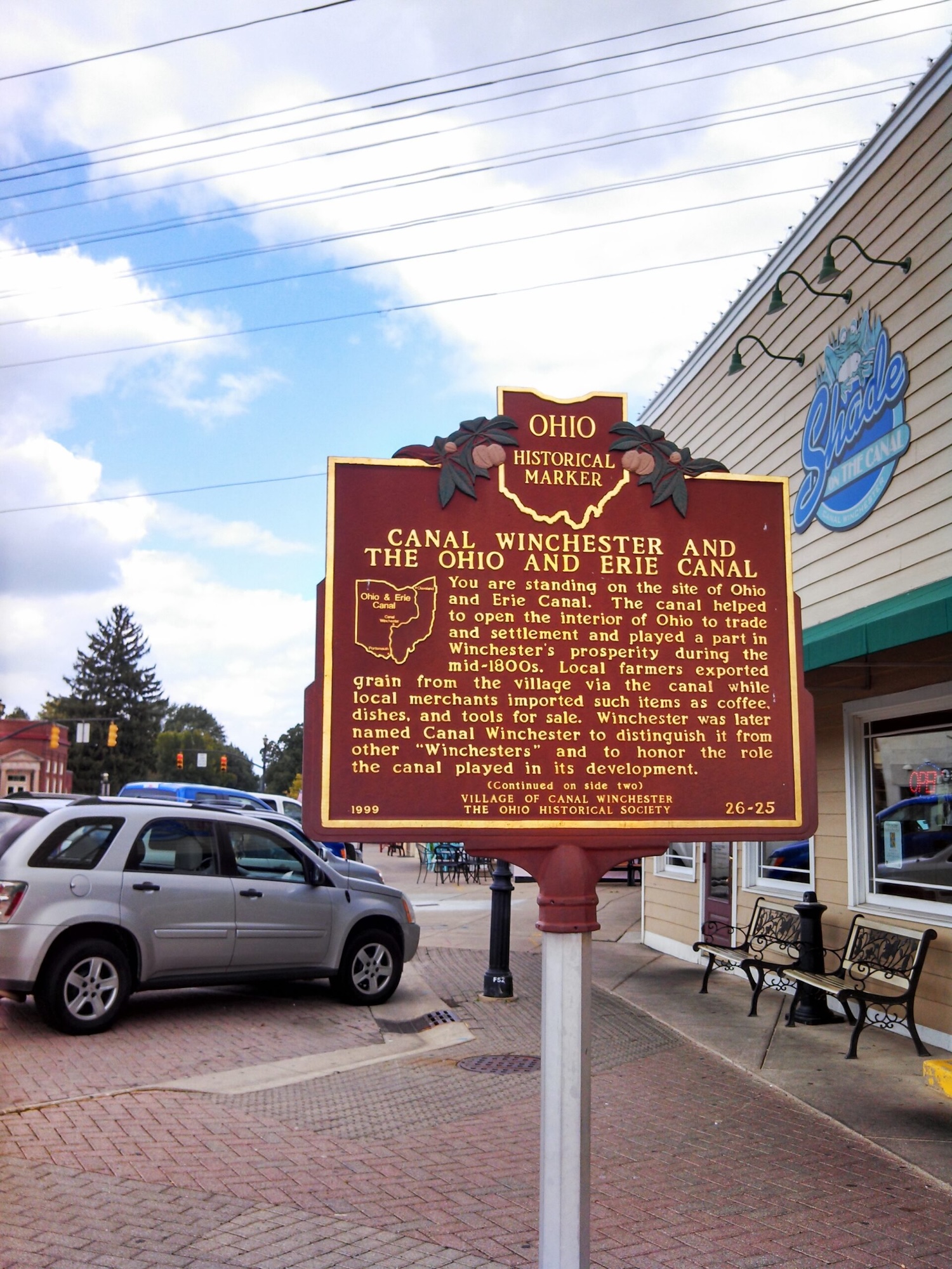 Ohio Historical Marker sign about Canal Winchester and the Ohio and Erie Canal on a street corner with cars and a cafe in the background.