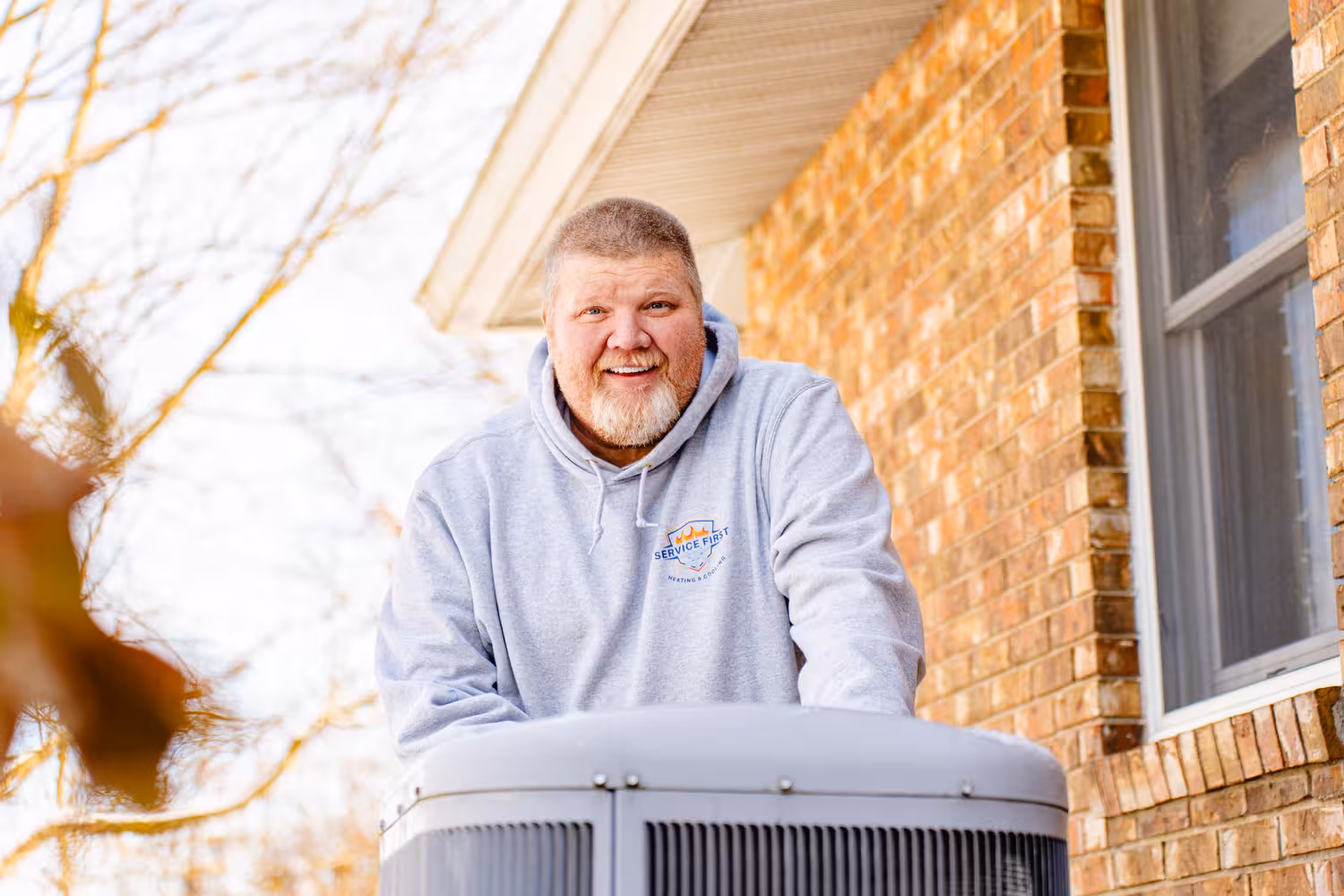 Smiling HVAC technician in a gray Service First hoodie standing behind an outdoor air conditioning unit by a brick house.