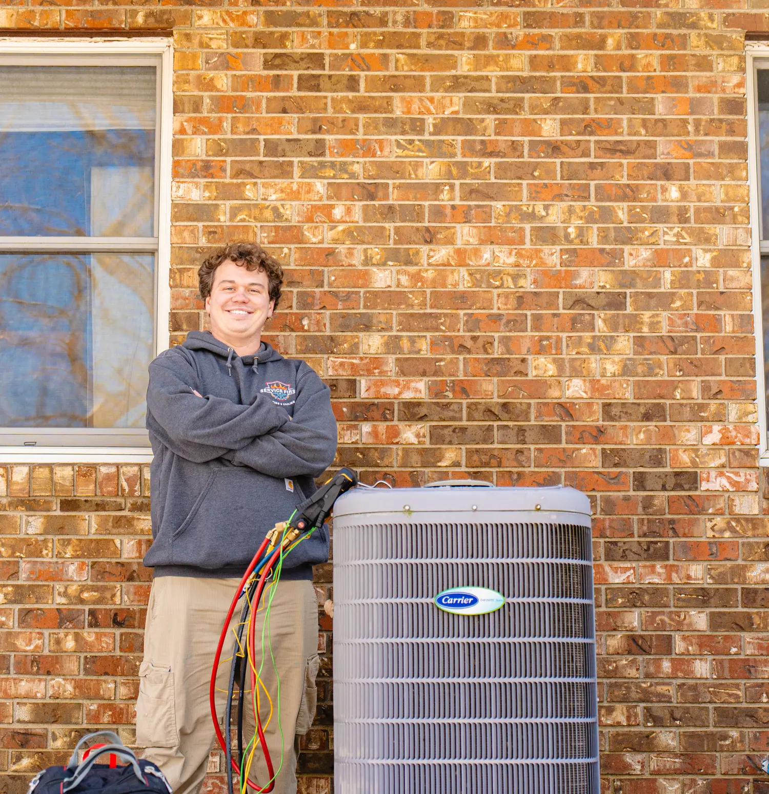 Technician in a gray hoodie standing next to a Carrier air conditioning unit against a brick wall.