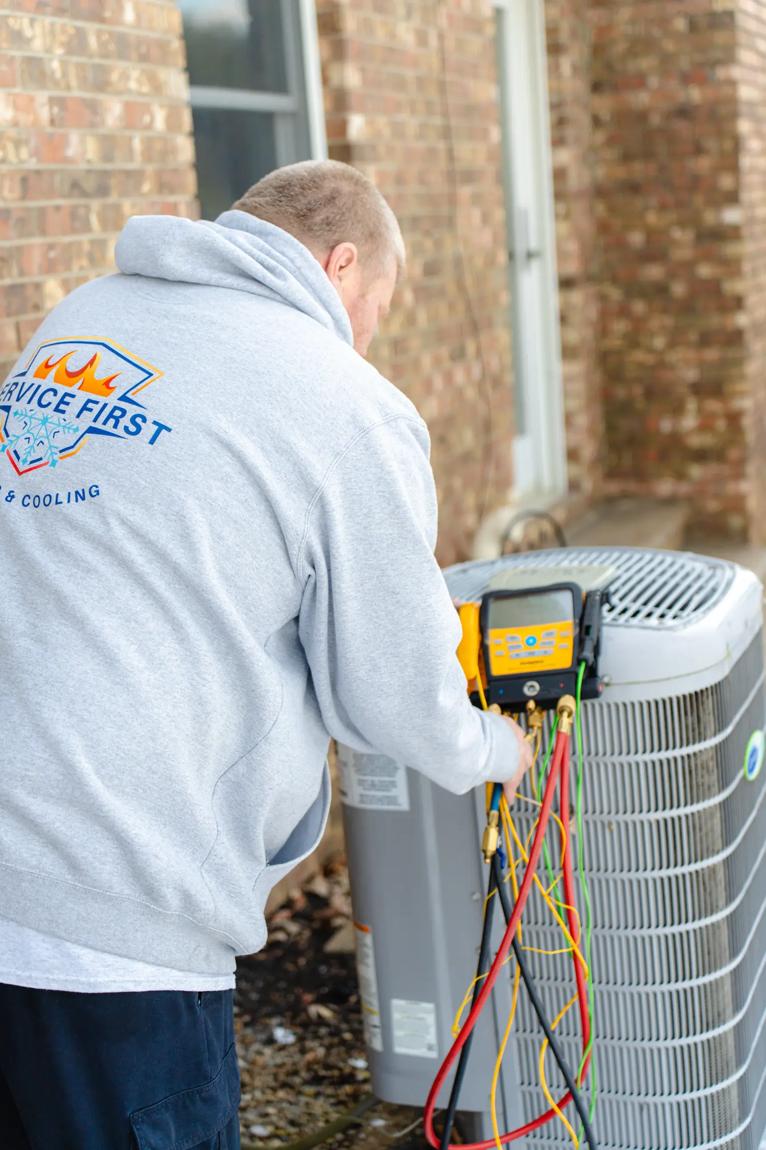 Technician wearing a gray hoodie inspecting an outdoor air conditioning unit with diagnostic tools and colorful wires attached.