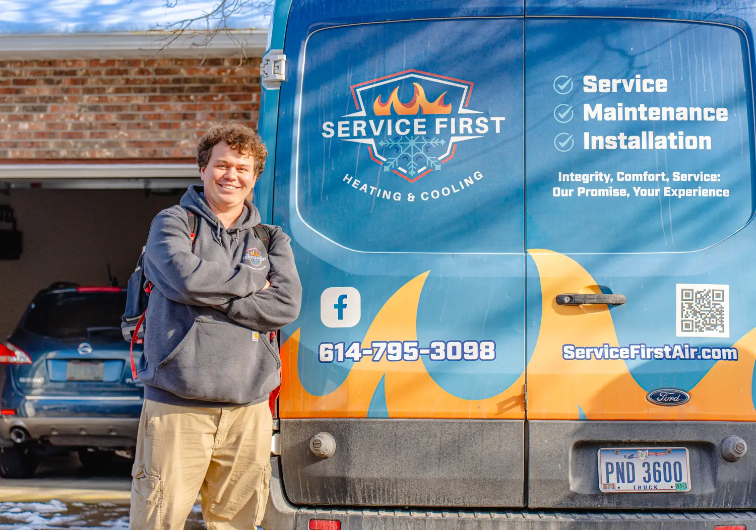 Man in a hoodie and cargo pants smiling with arms crossed next to a blue service van with flames and contact info for Service First Heating & Cooling in Lancaster, Ohio.