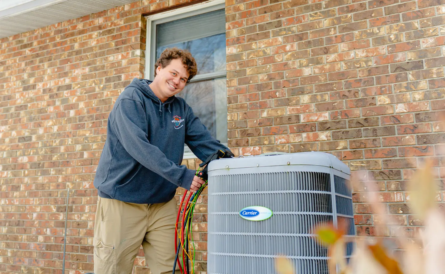 Technician smiling while working on an outdoor air conditioning unit beside a brick house wall in Lancaster, Ohio.
