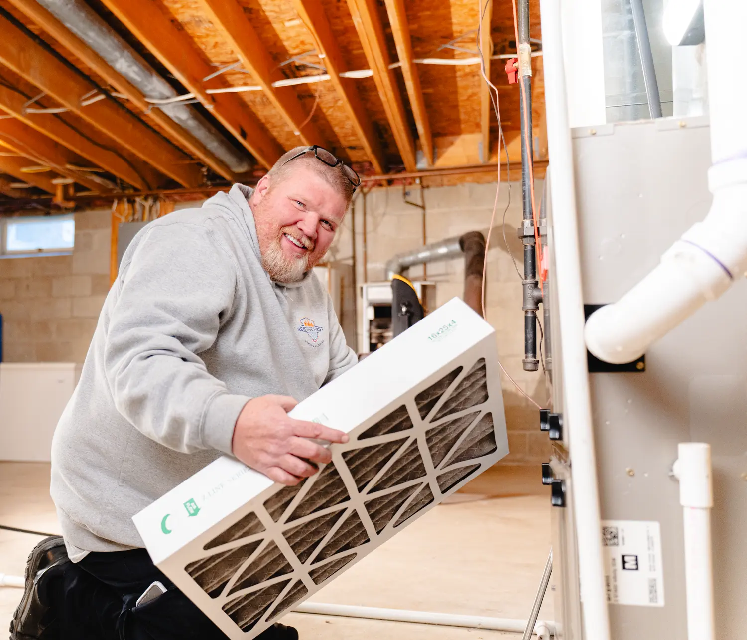Technician kneeling and holding a furnace air filter in a basement with exposed wooden ceiling beams in Lancaster, Ohio.
