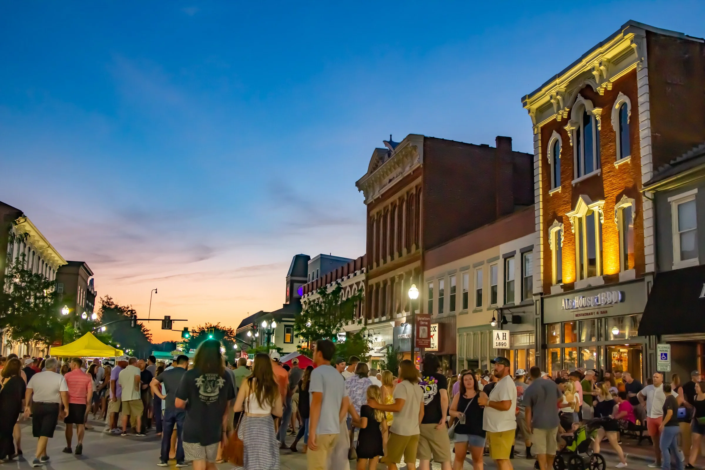 Crowd of people walking and socializing on downtown lancaster street at dusk with historic brick buildings and a restaurant lit up.