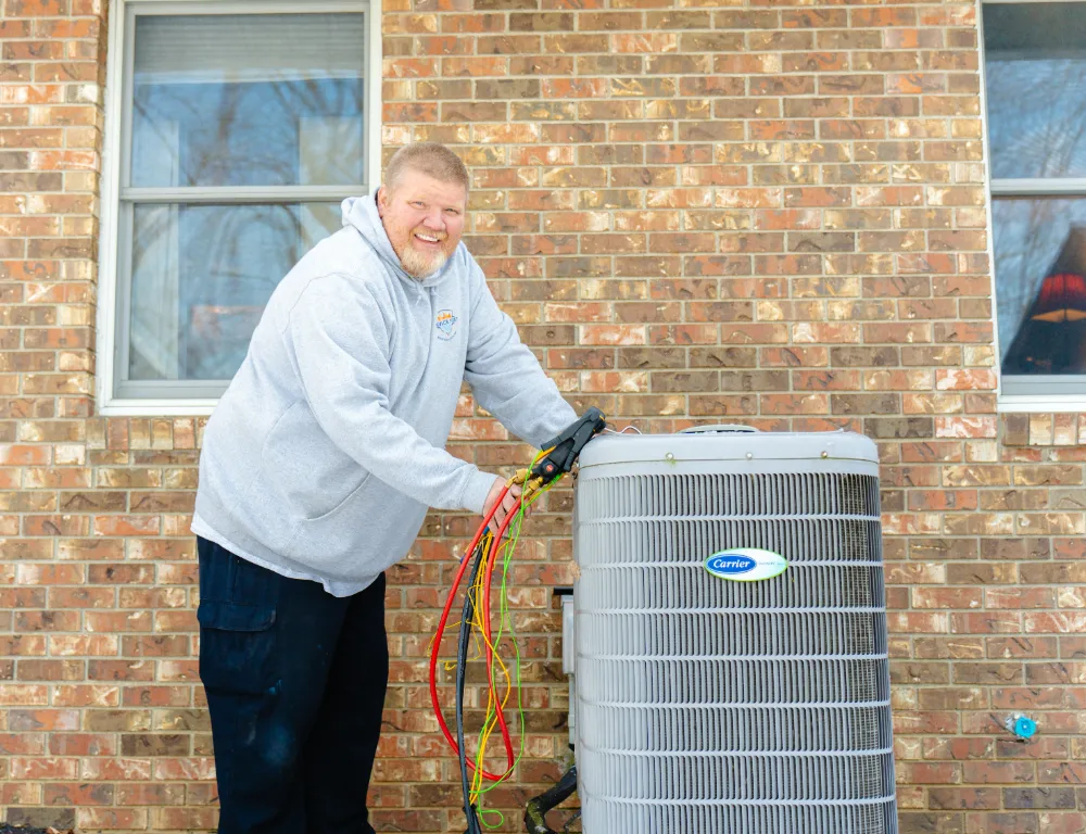 A man in a gray hoodie working on a Carrier air conditioning unit outside a brick house in Groveport.
