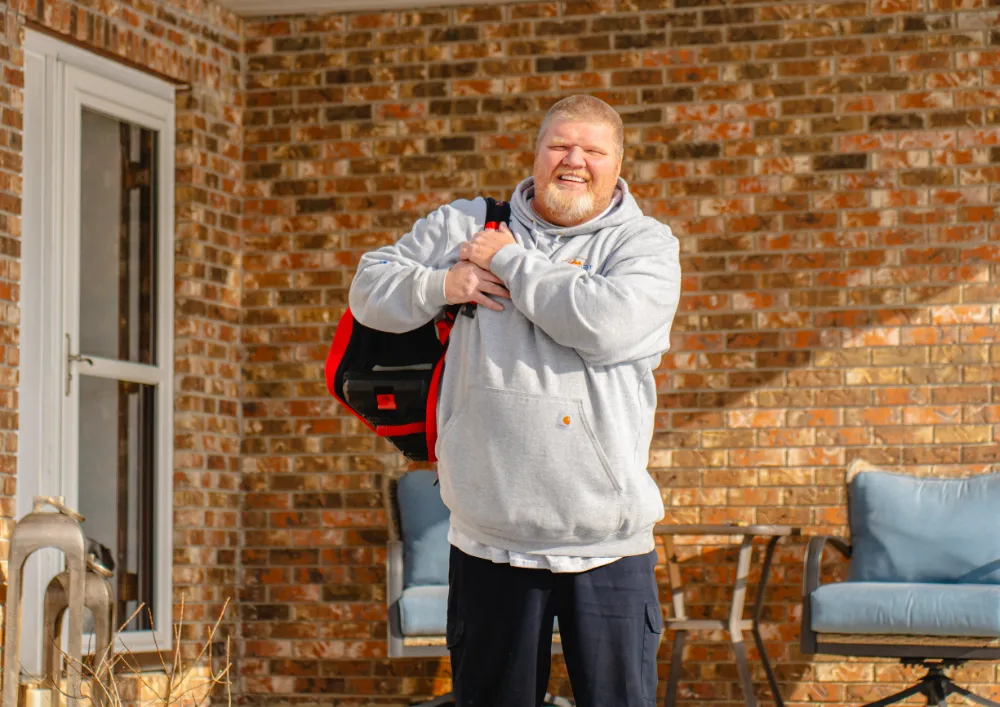Smiling man in a gray hoodie carrying a red and black bag standing outside a brick house with patio furniture in Reynoldsburg, Ohio.