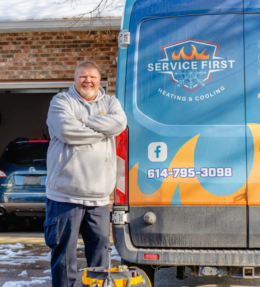 Smiling man in gray hoodie standing with arms crossed beside a blue heating and cooling service van in Reynoldsburg, Ohio.