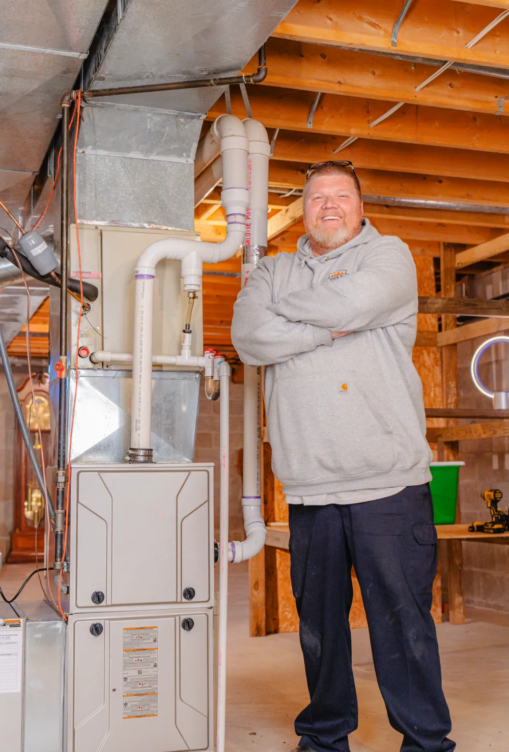 Man in gray hoodie and dark pants standing with arms crossed next to a home HVAC furnace system in a basement in Reynoldsburg, Ohio.