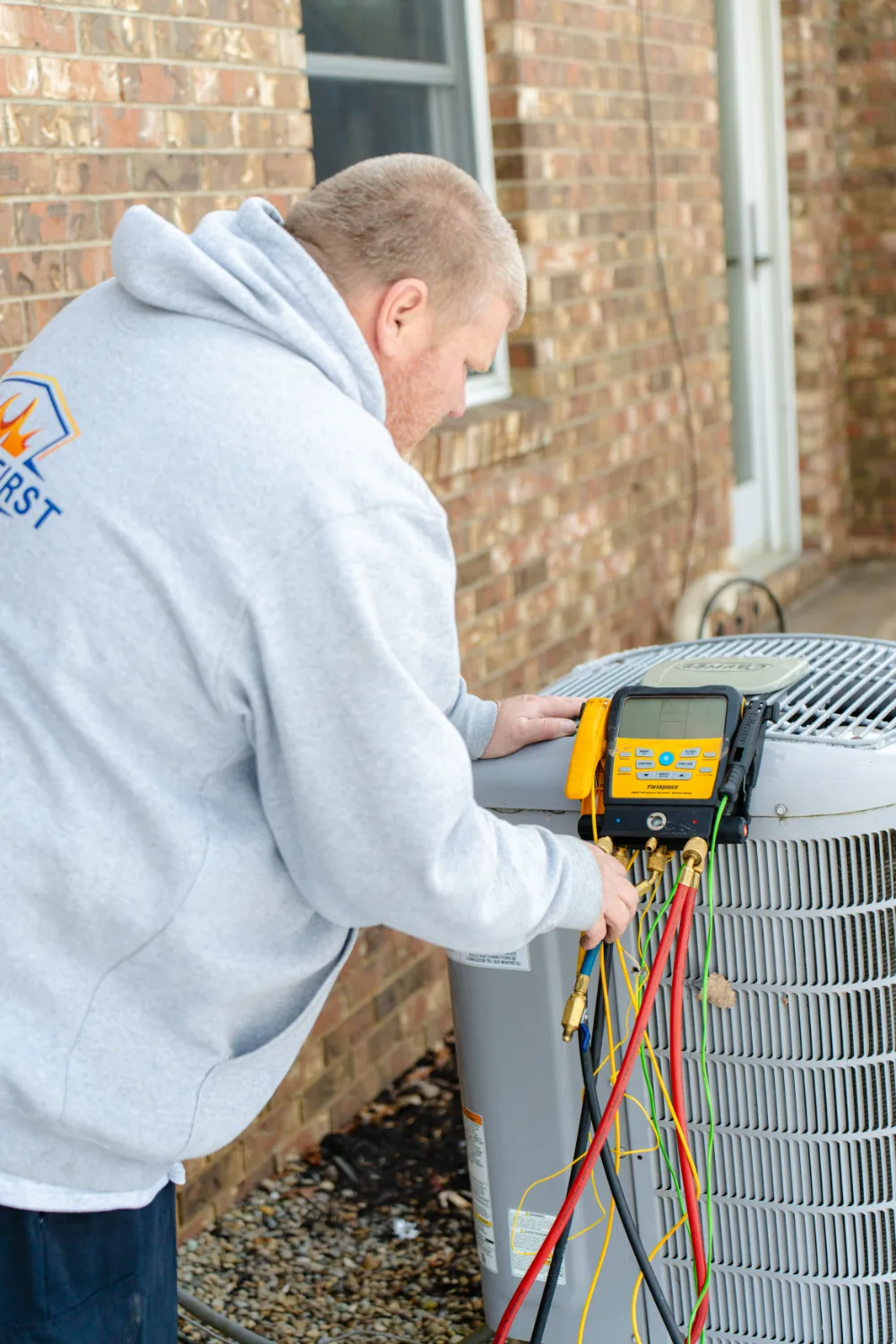 Technician in gray hoodie checks an air conditioning unit with diagnostic tools and colorful wires outdoors.
