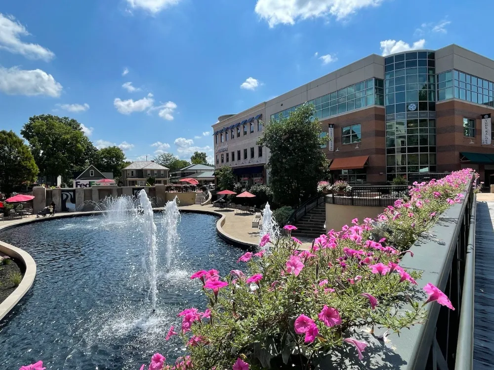 Outdoor fountain with water jets surrounded by pink flowers and buildings under a blue sky.