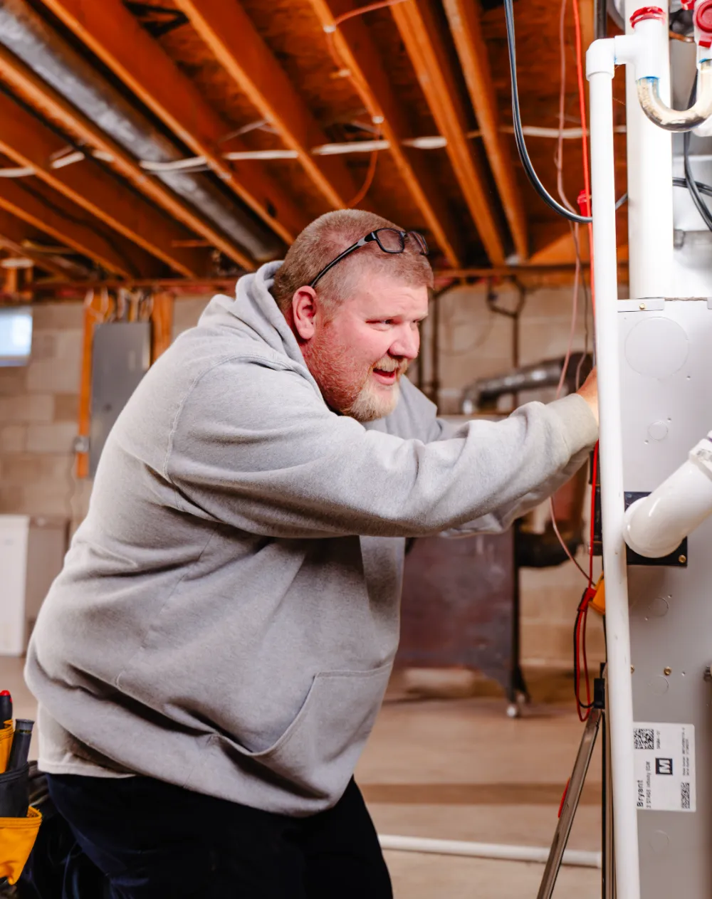 Technician in gray hoodie repairing furnace unit in basement with exposed ceiling beams.