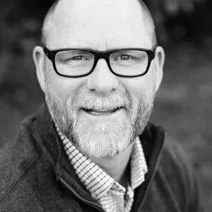 Smiling middle-aged man with glasses, a beard, and a collared shirt under a dark jacket.