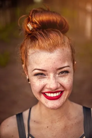 Smiling young woman with red hair in a bun, freckles, and bright red lipstick wearing a dark sleeveless top.