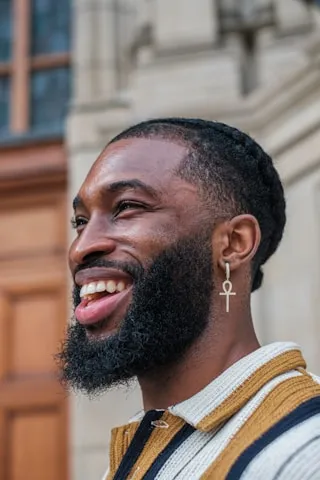 Smiling man with braided hair, full beard, and ankh earring wearing a striped shirt in front of a stone building.