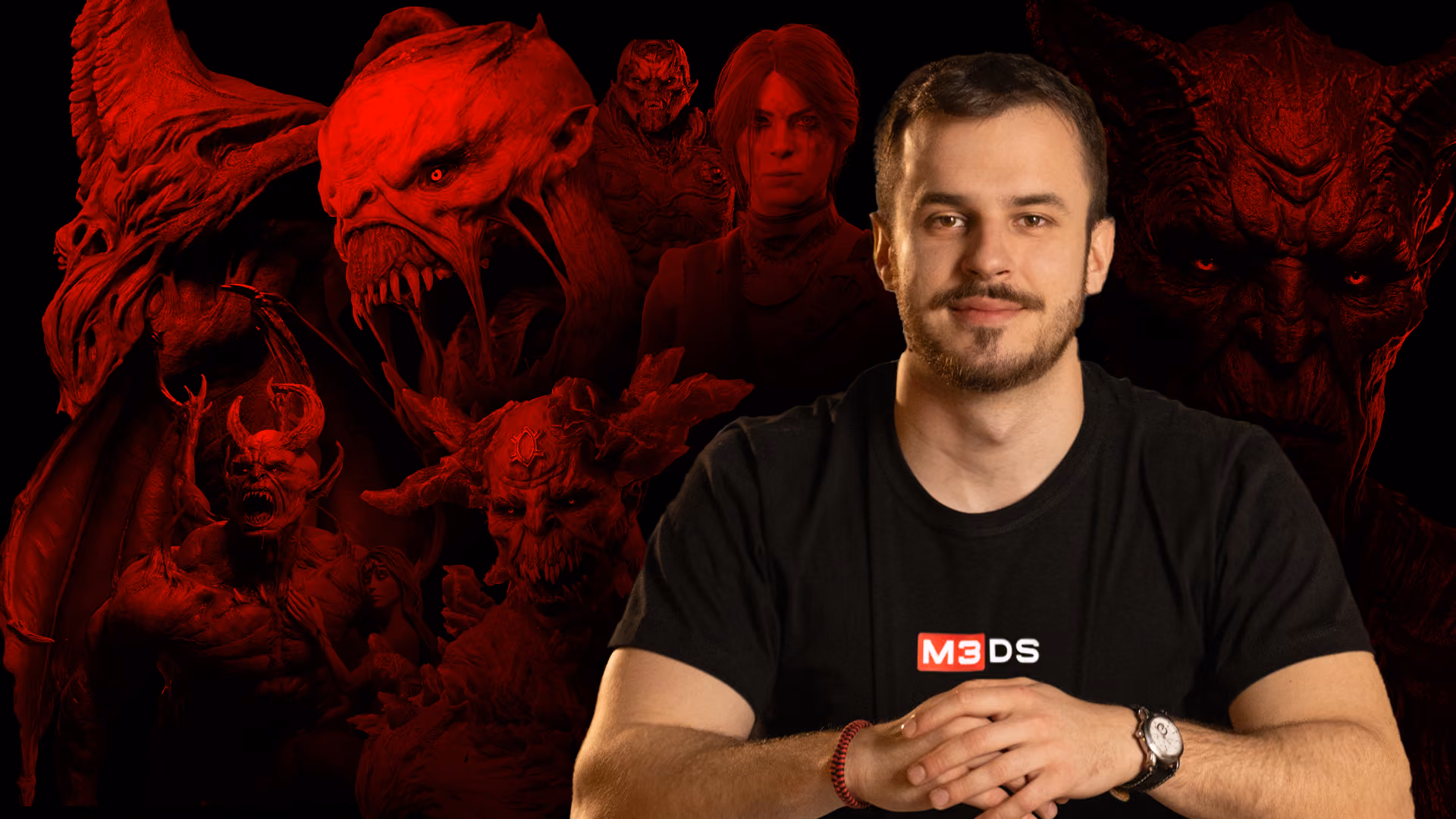 Man in black M3DS shirt sitting with hands clasped, red-lit demonic figures in the dark background.