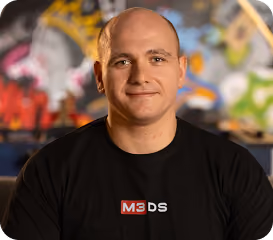 Smiling bald man wearing a black M3DS shirt with colorful blurred background.