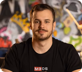 Smiling man with short hair and beard wearing black M3DS T-shirt in front of colorful graffiti wall.
