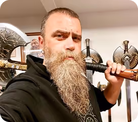 Bearded man holding a decorated battle axe over his shoulder, with shields displayed on the wall behind him.