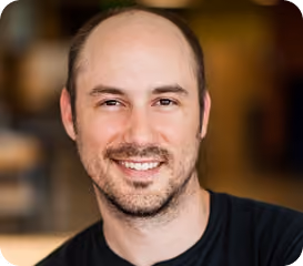 Smiling man with short beard and balding head wearing a black shirt with blurred background.