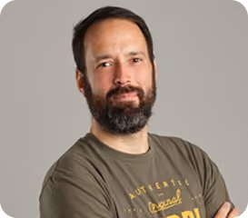 Bearded man with crossed arms wearing a green t-shirt against a plain gray background.