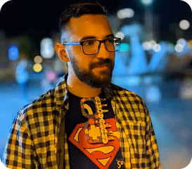 Man with glasses and beard wearing Superman shirt and checkered jacket at night outdoors.
