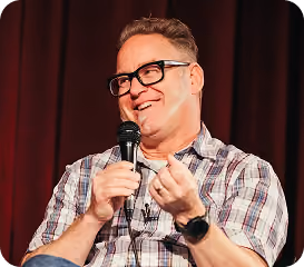 Smiling man in glasses and plaid shirt speaking into a microphone on stage with red curtains.
