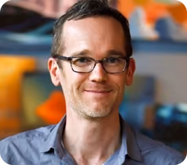 Smiling man with glasses and short hair wearing a blue collared shirt in a colorful indoor setting.
