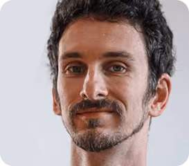Headshot of a man with dark curly hair, beard, and mustache, smiling slightly against a light background.