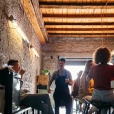 Serveur debout dans un café rustique avec clients assis et lumière naturelle entrant par une grande fenêtre.