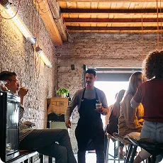Serveur debout dans un café rustique avec clients assis et lumière naturelle entrant par une grande fenêtre.