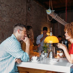 Un homme et une femme discutent en buvant au bar d'un restaurant animé avec un serveur en arrière-plan.