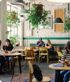 Intérieur d'un café avec des clients assis aux tables et de nombreuses plantes suspendues au plafond.